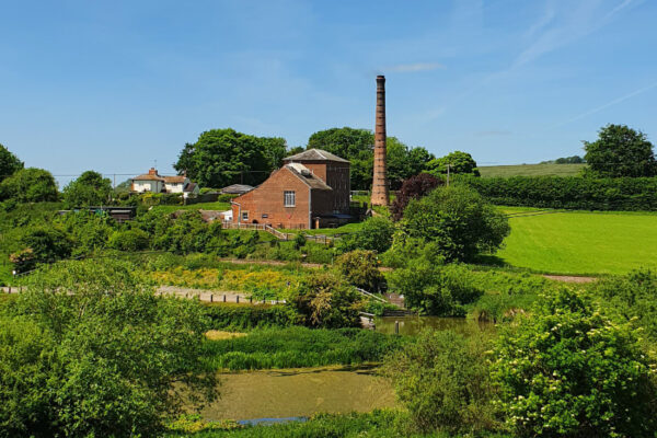 Terracotta brick building with tall brick chimney surrounded by green lush grass, trees and fields.