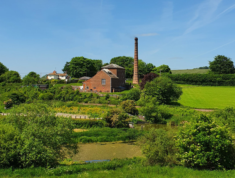 Terracotta brick building with tall brick chimney surrounded by green lush grass, trees and fields.