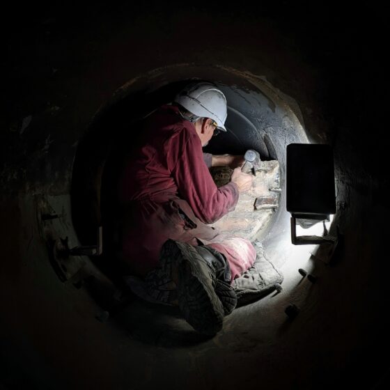 Figure in a burgundy boiler suit within a dark furnace tube 32 inches wide. A torch lights the space as they hammer at a brick wall within the tube.