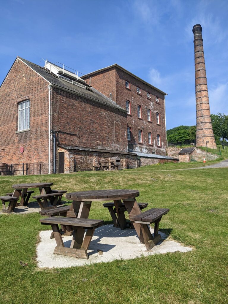 Round dark wood picnic tables cover a green lawn in foreground. A terracotta brick building towers behind, with 12 windows on one side and a 25 metre chimney. All backed by blue sky.